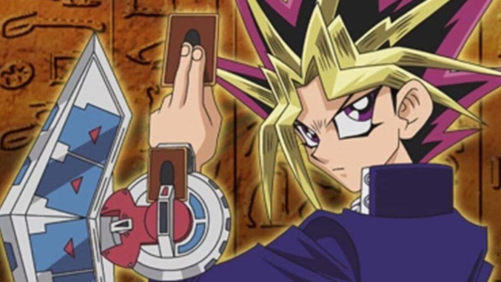 Yugi tenant une carte et montrant son disque de duel devant un mur recouvert de hiéroglyphes dans l'anime Yu-Gi-Oh